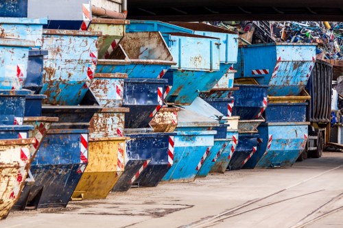 A row of large metal skip bins lined up along a paved area, with some showing signs of rust and wear. The bins are painted in shades of blue, with red and white striped reflective safety tape on the edges. They are arranged side by side, with their open tops visible, some partially filled with mixed waste materials. The surrounding environment appears to be an industrial or commercial waste collection site, with a dark truck or container partially visible on the right. The ground beneath the skips is marked with tire tracks and dirt, suggesting frequent use for rubbish disposal and waste removal activities in the Vauxhall area. Commercial Waste Vauxhall’s service often involves handling such large bins for debris, construction waste, or general rubbish from nearby commercial premises.