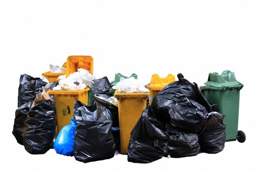 A collection of black, blue, and yellow rubbish bags filled with waste is stacked on a paved driveway or pavement area in front of a row of four wheelie bins, each a different colour: yellow, green, and additional black in the background. The bags appear to contain domestic or commercial waste, with some partially opened revealing paper and plastic materials. The wheelie bins are made of durable plastic with smooth surfaces and are positioned side by side, with the yellow bin slightly overlapping the green one. The scene suggests a waste collection or rubbish removal operation in an urban or suburban environment, possibly in Vauxhall or nearby London postcodes, managed by Commercial Waste Vauxhall for a commercial or property premises. The background is plain white, emphasizing the waste and containers. The image highlights waste management efforts typical of local rubbish removal services, with a focus on the segregation and disposal of different types of waste on residential or commercial property driveways or frontages.