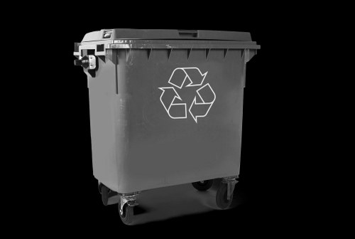 A large, black plastic waste bin with a hinged lid and four wheels, positioned outdoors on a paved surface. The bin features a white recycling symbol on its front, indicating it is used for recyclable rubbish. The surface of the bin appears textured, with visible ridges along the top edge and a slightly glossy finish. The background is plain and dark, isolating the bin as the main subject. The bin's positioning suggests it is ready for collection or disposal, fitting within the context of commercial waste management services in Vauxhall, London. The image does not show the contents inside or any surrounding environment, focusing solely on the waste container, which aligns with the rubbish removal services offered by Commercial Waste Vauxhall.