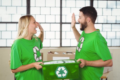 A photograph shows two young adults, a woman and a man, standing indoors in a brightly lit space with large industrial-style windows and a brick wall background. Both are wearing bright green t-shirts featuring the recycling symbol. The woman has blonde hair and is smiling broadly, with her right hand raised to her face as if laughing or expressing surprise. The man has short dark hair and a beard, and is smiling as well, raising his right hand for a high-five. Between them, the man is holding a transparent green recycling bin with a white recycling symbol on its side, positioned at waist level. The environment suggests a clean, organized space, possibly a waste management or recycling facility, or an office involved in sustainability efforts. This image relates to environmentally conscious disposal practices and, by extension, to rubbish removal services like those provided by Commercial Waste Vauxhall, especially emphasizing recycling and responsible waste handling within the Vauxhall area.